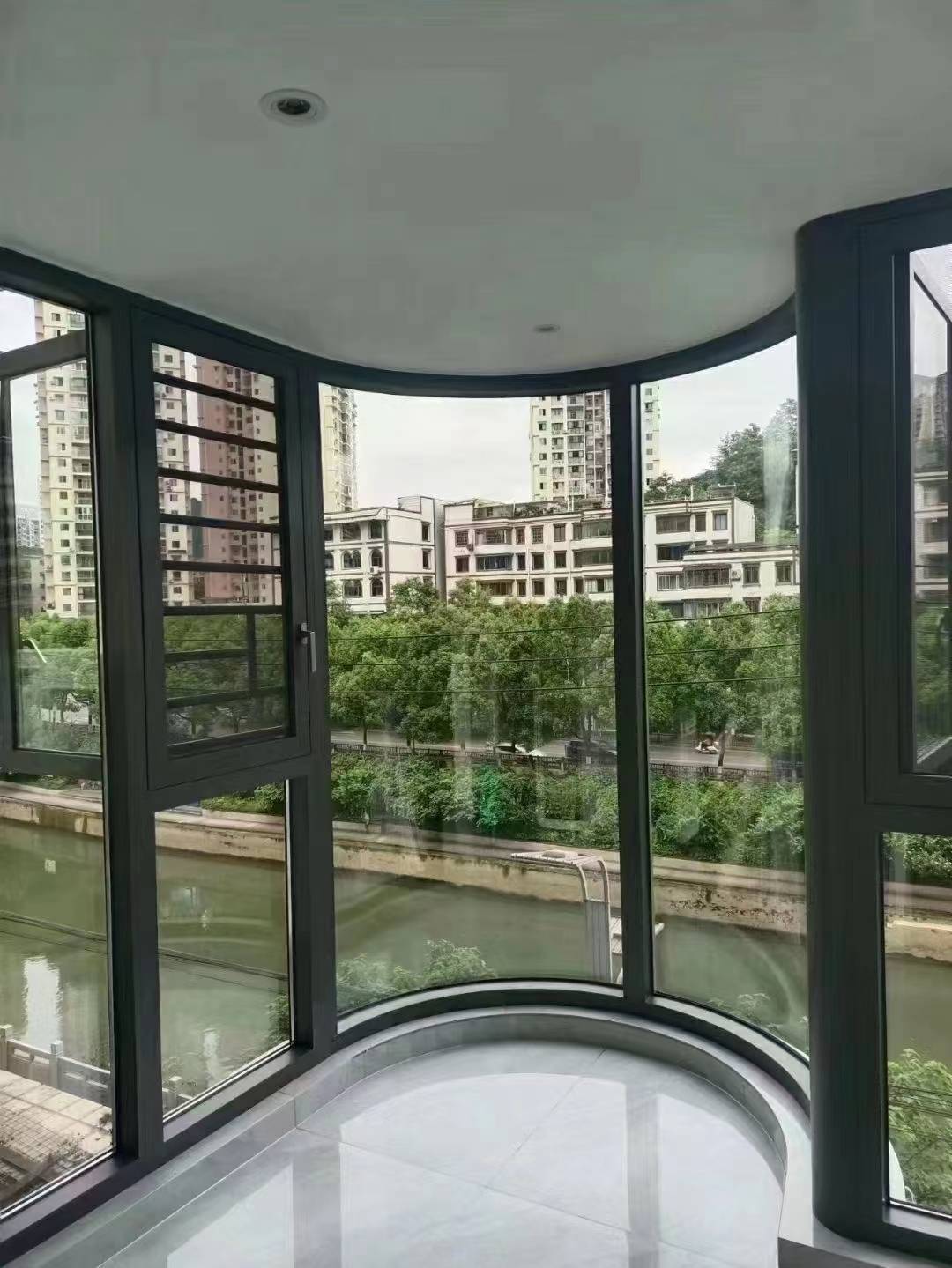 现代简约风格的弧形阳台封窗实拍。深灰色断桥铝窗框搭配高透玻璃,呈流畅圆弧造型,完美贴合空间结构。左侧开启扇配备金刚网纱窗,兼顾通风防蚊。窗外临河绿树景观一览无余