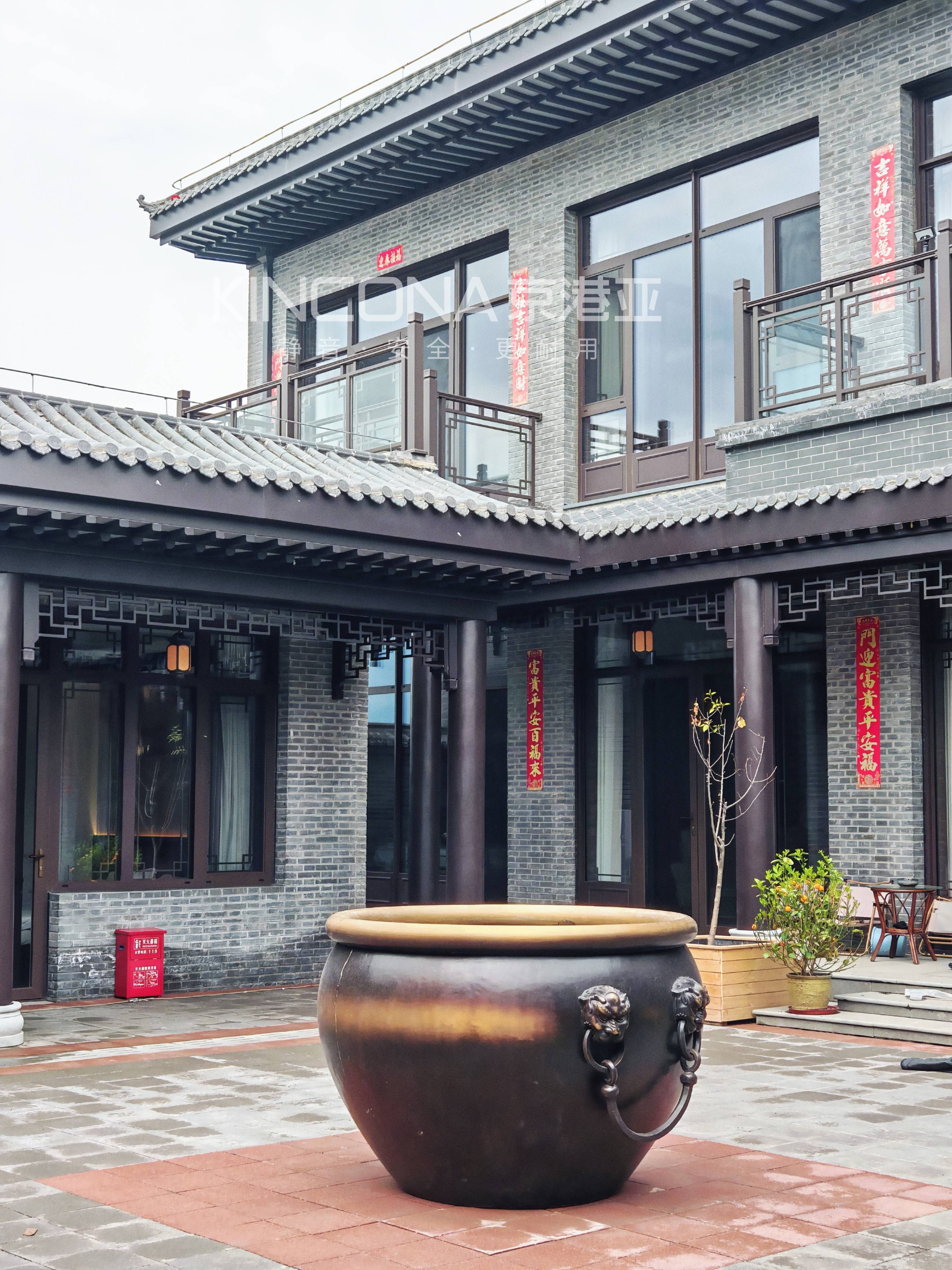 这是一张新中式庭院的实景照片,主体为一座两层青砖灰瓦的别墅建筑,前景摆放着巨大的兽首铜缸。建筑安装了深棕色的京港亚系统门窗,搭配中式回纹栏杆,展现了传统美学与现