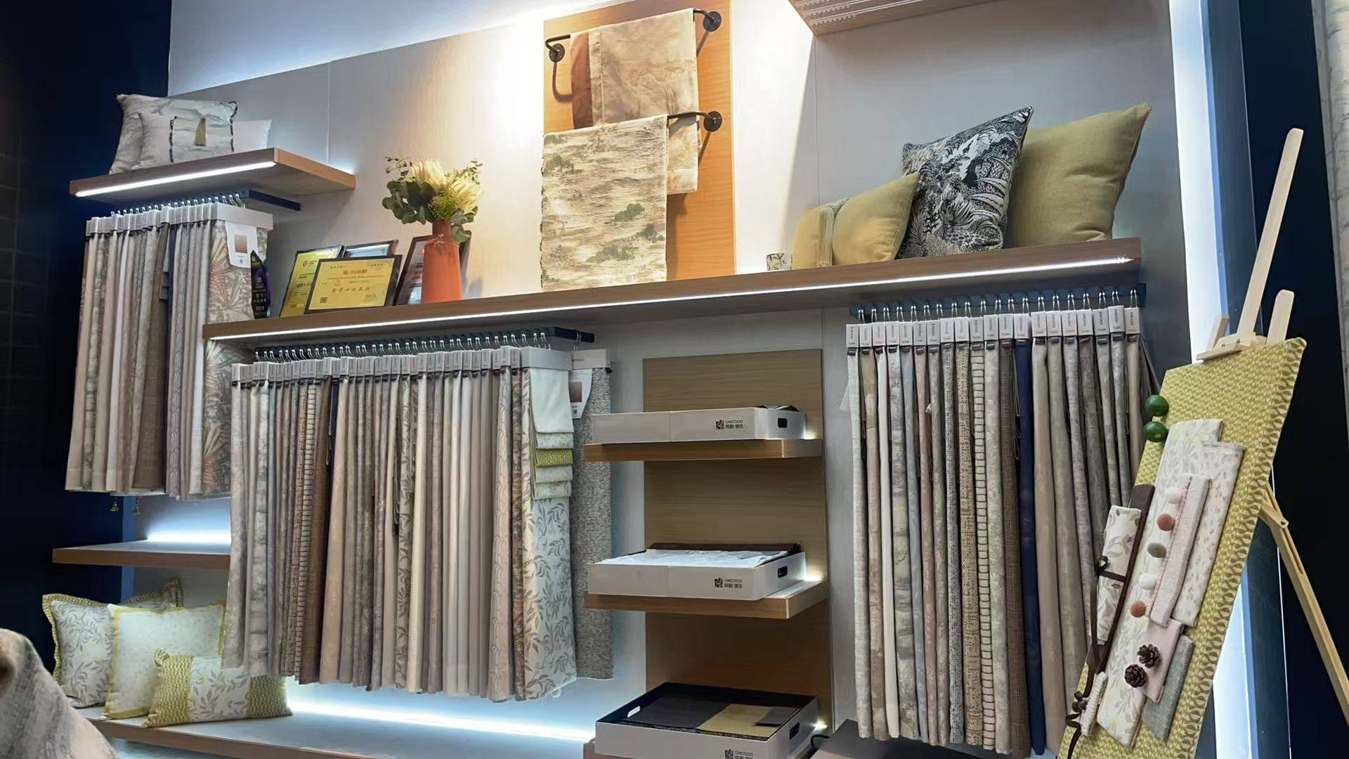 家居软装展厅内，多排窗帘面料样品整齐悬挂，涵盖米灰绿等自然色系及印花纹理。木质搁板搭配抱枕与装饰画架，展示丰富材质细节。暖色灯带营造温馨氛围，直观呈现布艺质感，